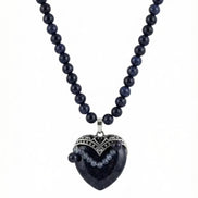 Colier pe fundal alb din mărgele albastre sclipitoare și pandantiv mare în formă de inimă cu montură argintie filigranată.