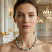 Femeie purtând un colier scurt la baza gâtului din pietre verzi fațetate și perlă alungită, asortat la o rochie crem cu umerii goi.