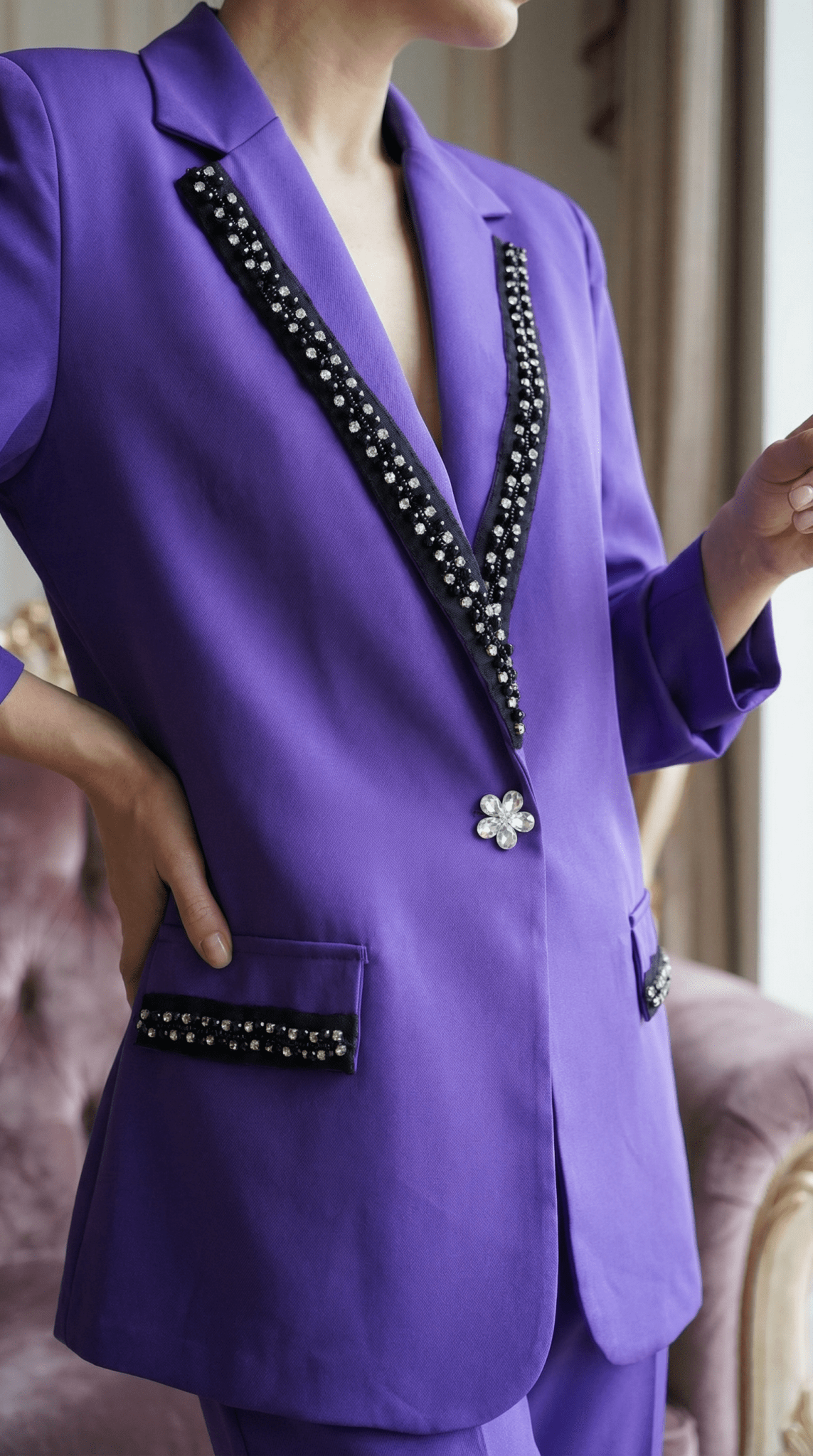 Vedere de aproape a buzunarului cu clapă neagră și cristale de pe un sacou mov și a nasturelui central strălucitor.