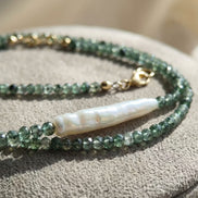 Prim-plan macro evidențiind textura neregulată a perlei centrale albe și fațetele mărgelelor verzi translucide pe fundal textil.