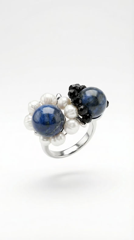 : Inel argintiu pe fundal alb cu montură voluminoasă din pietre albastre sferice perle albe neregulate și mărgele negre fațetate.