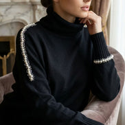 Femeie așezată purtând o maletă neagră cu guler înalt decorată cu perle pe linia umerilor și la manșete într-un decor interior luminos.