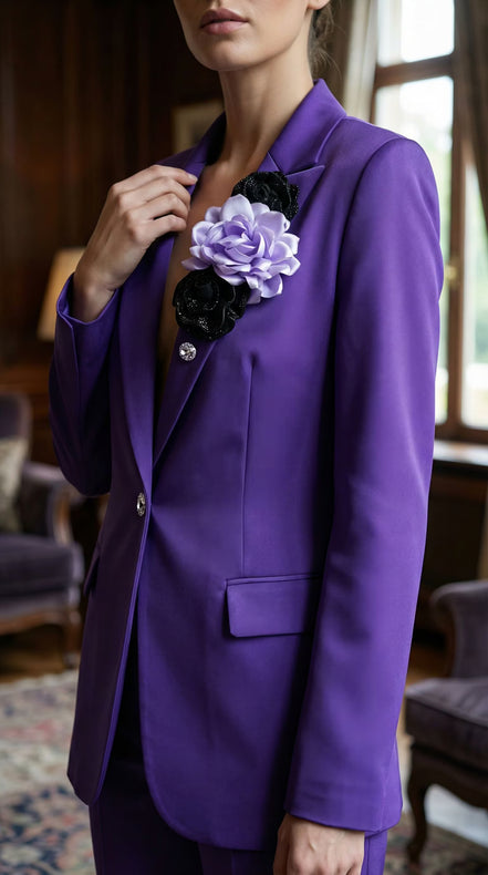 Femeie purtând un sacou mov structurat decorat pe rever cu un trandafir satinat lila și flori negre asortat cu nasture bijuterie.