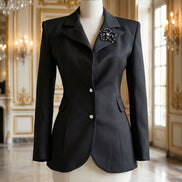 Sacou negru cambrat pozat pe manechin de croitorie cu revere ascuțite satinate nasturi triunghiulari și broșă florală aplicată.