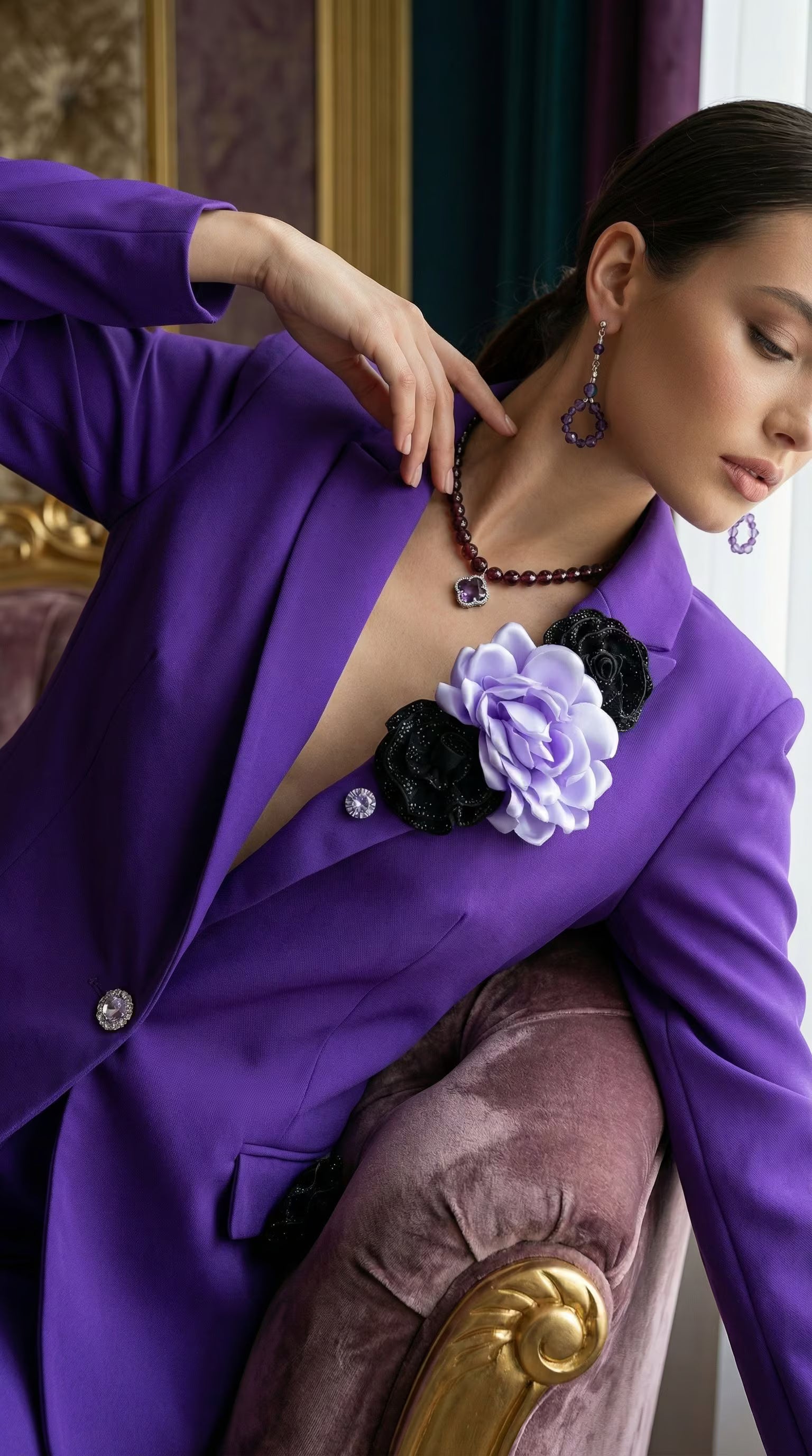 Model într-o ținută elegantă purtând sacou mov cu decorațiuni florale asortat cu cercei și colier din pietre mov într-un decor clasic.